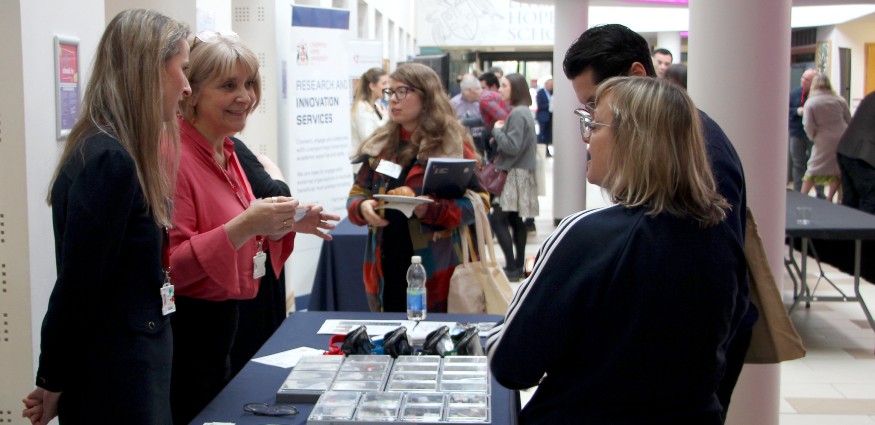 Two Caucasian females standing behind a desk with leaflets on speak to a Caucasian male and female.